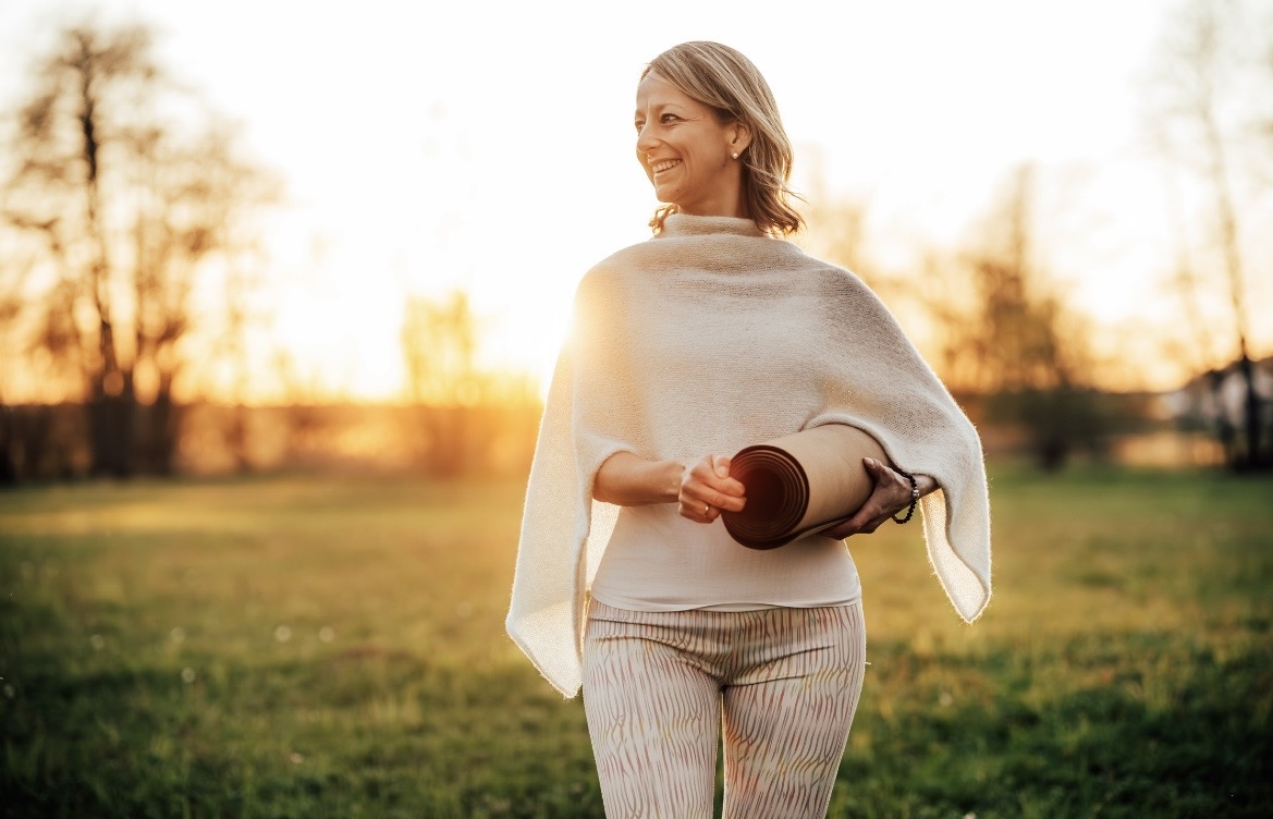Hanna Feld lächelt auf einer Wiese im Abendlicht, die Yogamatte unter dem Arm